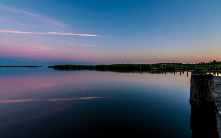 A very still body of water reflects the pink, purple and blue sky above. There is a dock in the foreground and trees lining the shore in the background. 